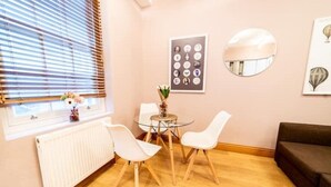 A room with a wooden floor, a white radiator, a window with blinds, a round glass table with two white chairs, a brown sofa, and wall art including a framed picture and a mirror. - St Paul Cathedral-City of London]London Apartment (London)