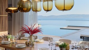 A dining area with a long table set for a meal, multiple hanging pendant lights, and a view of the sea. - Villa Mirabel, VILLA MIRABEL 5* (Mimice)