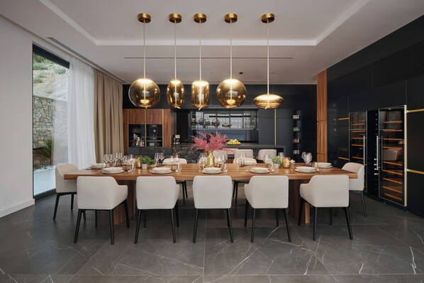 A modern dining room with a long table set for six, surrounded by white chairs. There are hanging gold pendant lights above the table.