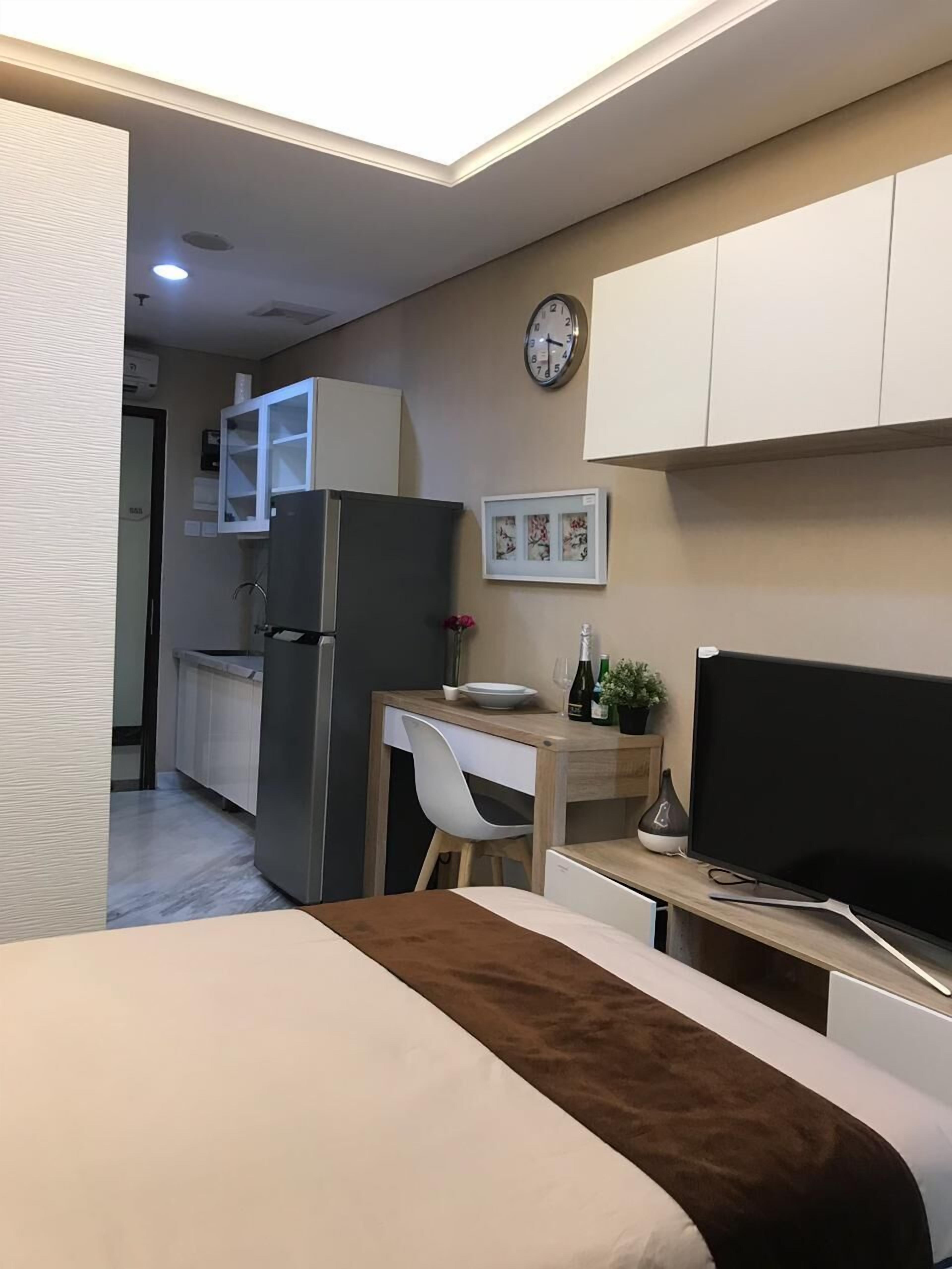 A compact hotel room with a bed, a desk with a chair, a television, a refrigerator, and a clock on the wall.
