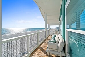 A balcony with a view of the beach and ocean, furnished with a white sofa and cushions, and a glass railing. - Constellation: Stunning 6 Bedroom New Build- Priv. Pool, Pet Friendly Sleeps 24! (Mexico Beach)