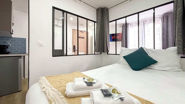 Une chambre d’hôtel moderne, équipée d’un lit, d’une table de chevet, d’un miroir et offrant une vue sur la ville grâce à de grandes fenêtres.