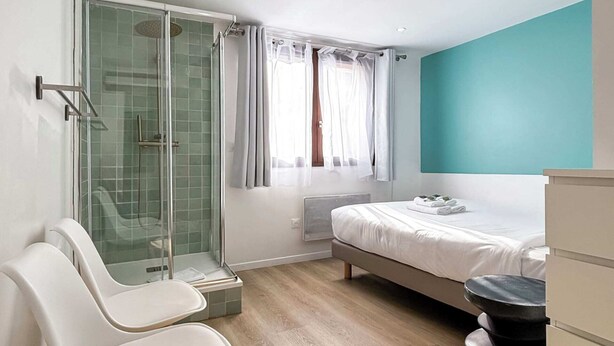 Un dormitorio moderno con cabina de ducha de vidrio, cama con ropa de cama blanca, pared de acento color turquesa y suelo de madera.