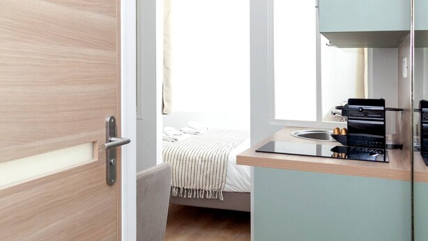 A compact kitchen with a sink, stove, and cabinets, a small trash bin, and a bed with a striped bedspread in the background. - David D Angers 26 - One Bedroom Apartment, Sleeps 2 (Paris)