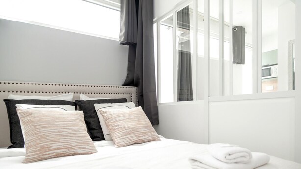Una cama bien hecha con sábanas blancas y dos almohadas, una ventana con cortinas y vistas a una zona de cocina.