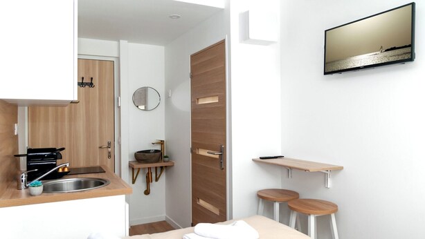 A modern hotel room with a wooden door, a round mirror, a sink, a small table, two stools, and a TV mounted on the wall. - David D Angers 22 - Studio Apartment, Sleeps 2 (Paris)