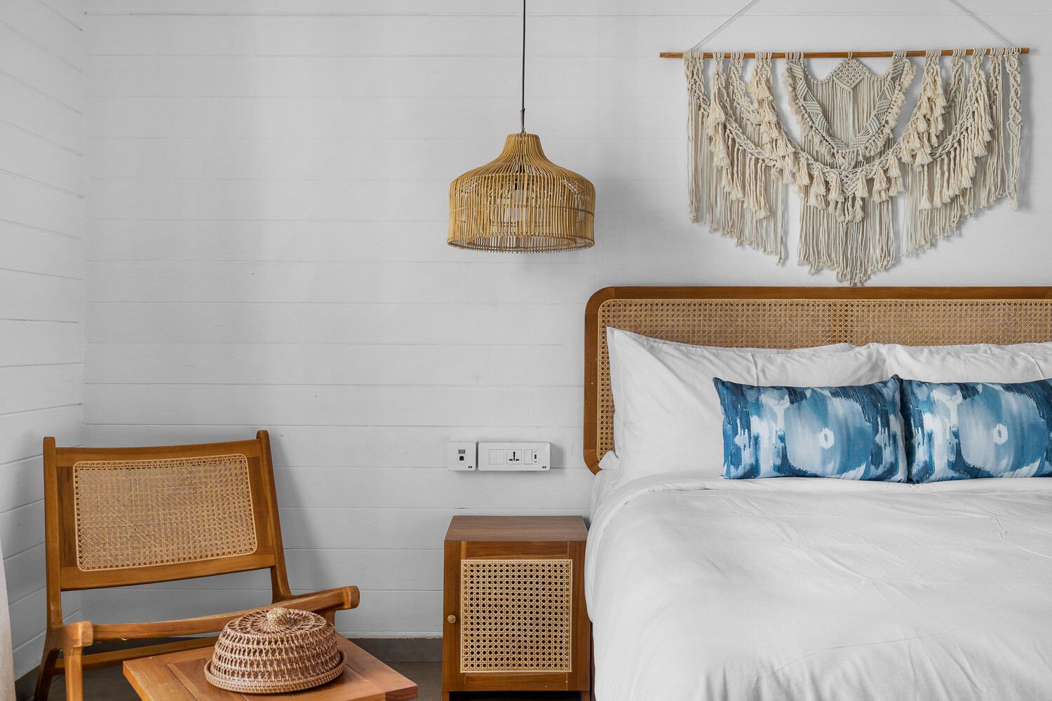 Ein Schlafzimmer mit Holzkopfteil, weißer Bettwäsche, einem gewebten Sessel, einem gewebten Nachttisch, einer gewebten Lampe und einem Wandbehang aus Makramee.