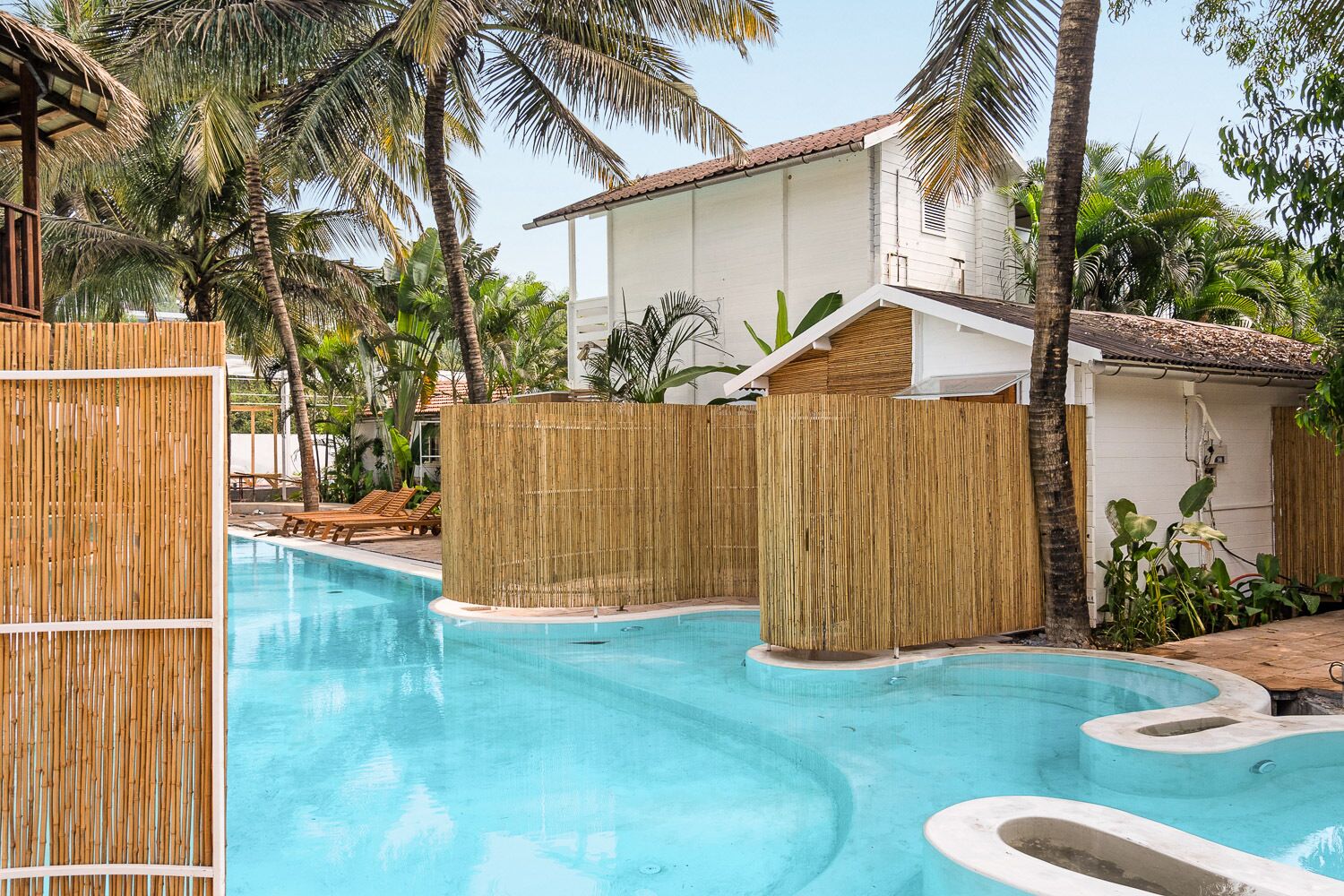 Un espace piscine agrémenté d’une clôture en bambou, de chaises longues et d’un bâtiment blanc en arrière-plan.