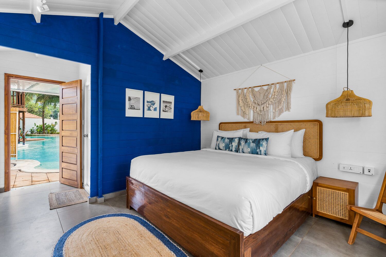 A bedroom with a large bed, a wooden headboard, a bedside table, a wall-mounted macrame piece, and a view of a swimming pool through an open door.