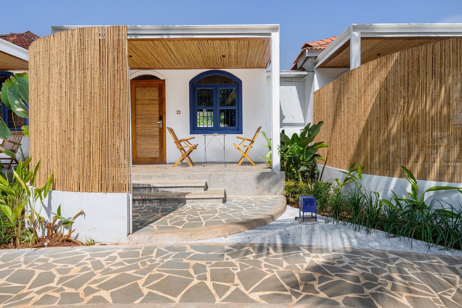 Une maison moderne avec une porte bleue, une porte en bois et une fenêtre bleue. Il y a deux chaises en bois à l’extérieur.