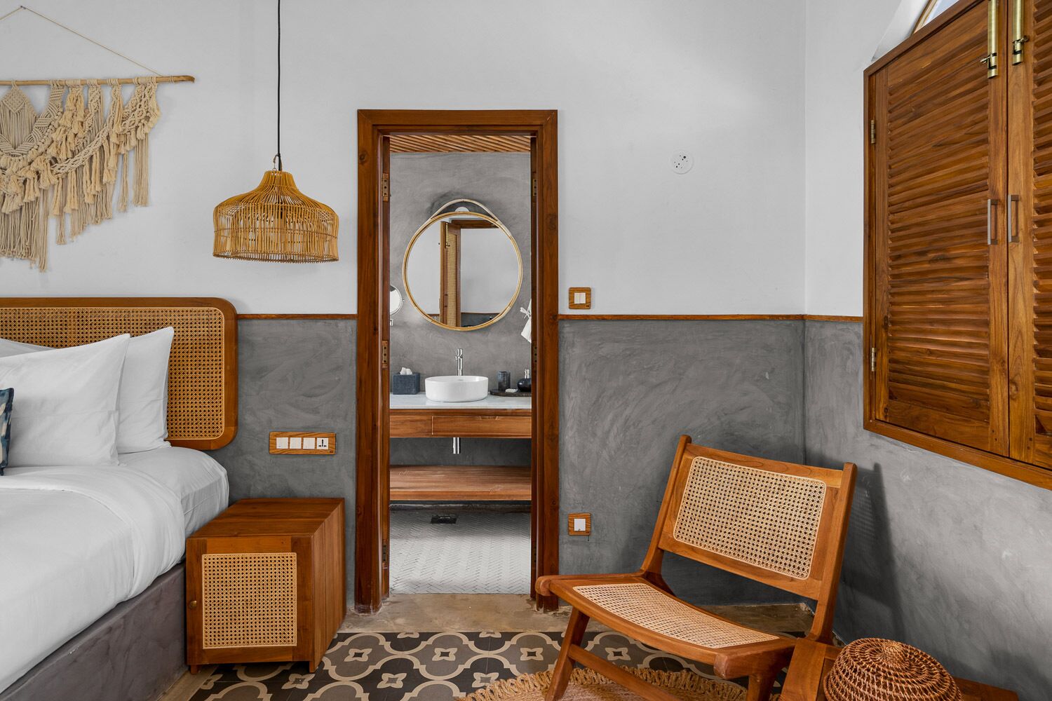A bedroom with a bed, bedside table, rattan chair, and a bathroom with a sink and mirror.