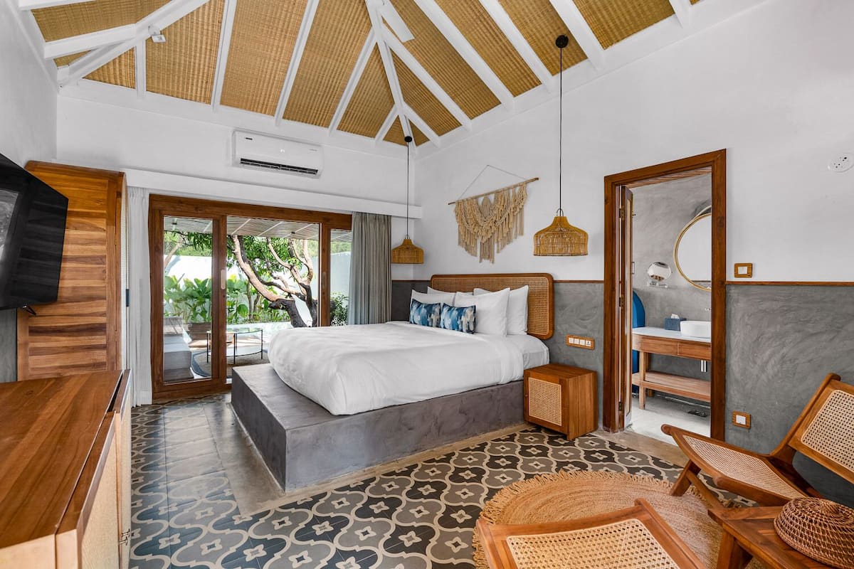 A modern bedroom with a large bed, a television, a bathroom with a mirror, and a view of greenery outside.