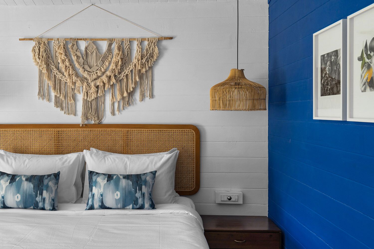 Un lit avec une literie blanche et des coussins à motifs bleus, une décoration murale en macramé, un luminaire suspendu en fibres tressées et des œuvres d’art encadrées sur un mur bleu.