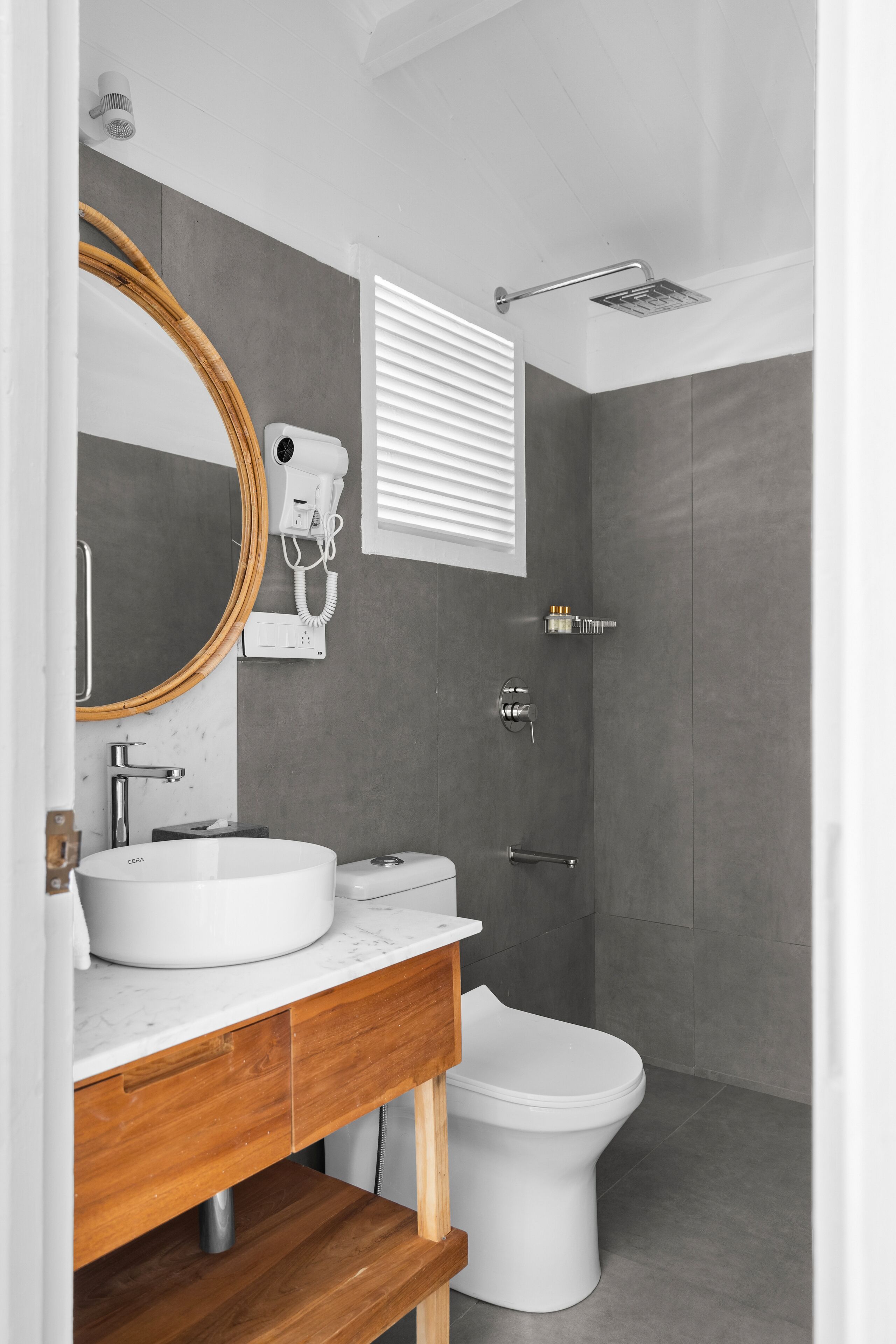 Un baño moderno con lavamanos blanco, tocador de madera, un espejo redondo, inodoro y zona de ducha.
