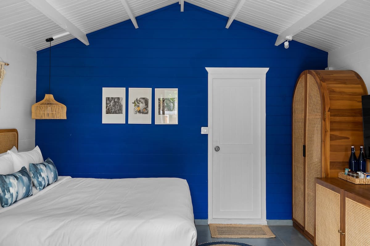 A bedroom with a bed, white bedding, a blue accent wall, a white door, and a wooden cabinet with bottles.