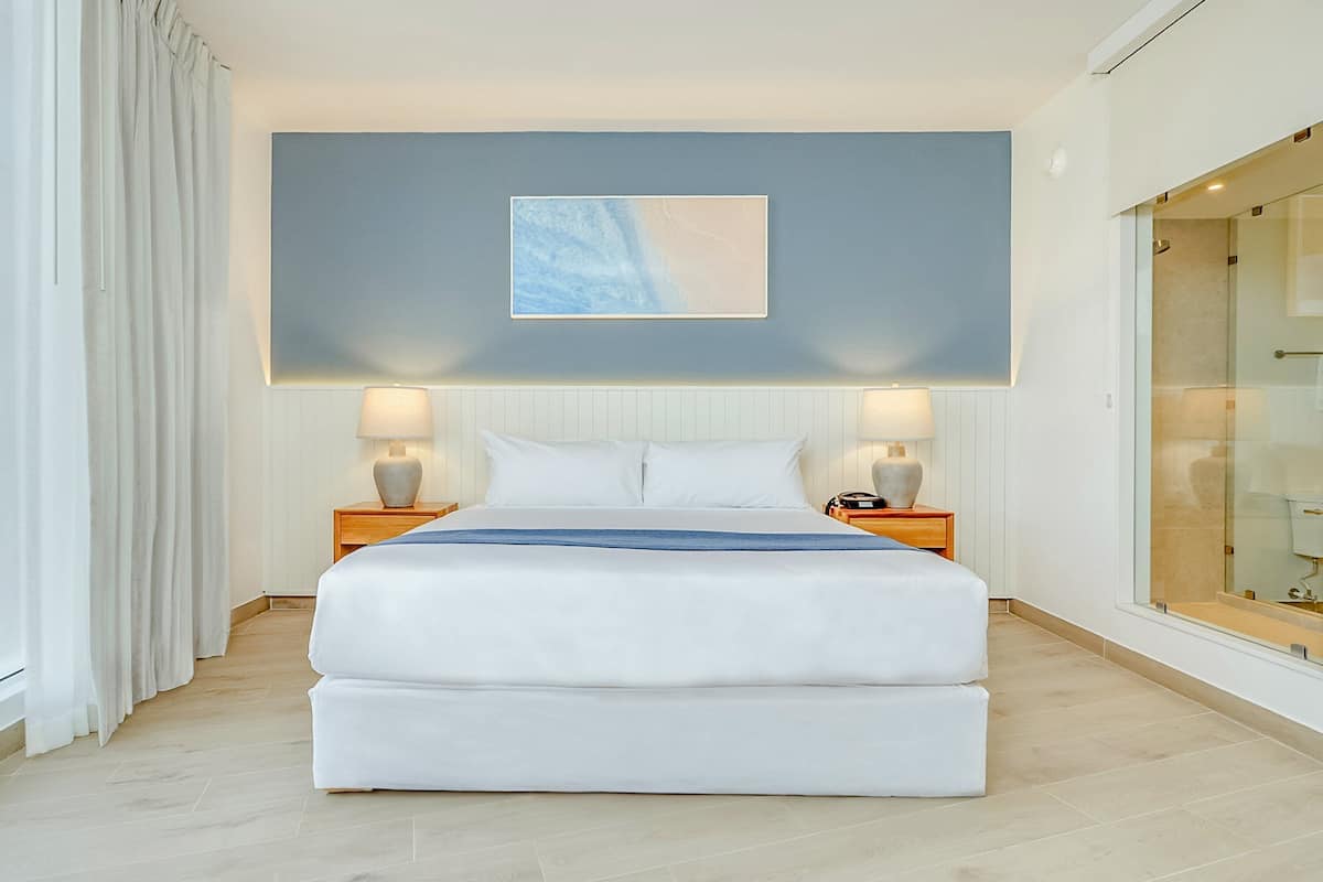 A modern hotel room with a large bed, two bedside tables, a wall-mounted artwork, and a glass shower enclosure.