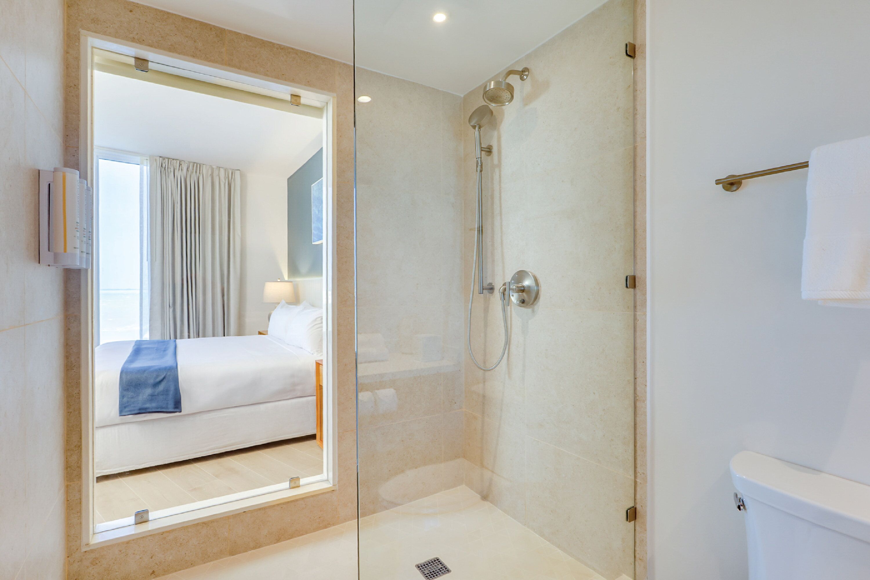 Ein modernes Badezimmer mit einer gläsernen Dusche, einem sichtbaren Bett mit blauer Decke und einer weißen Toilette.