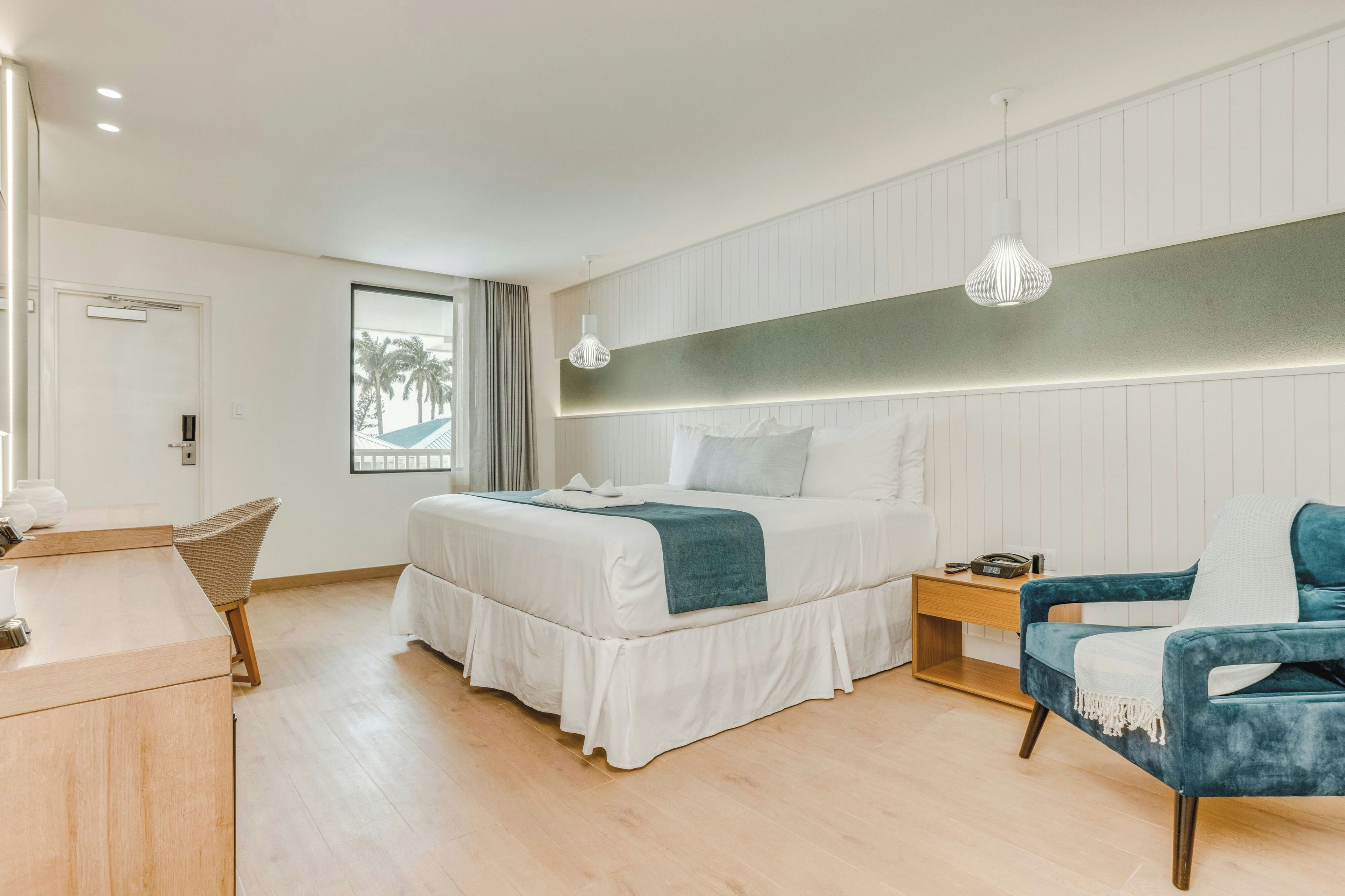 Una habitación de hotel moderna con una cama grande, un escritorio, una silla y vista a palmeras a través de la ventana.