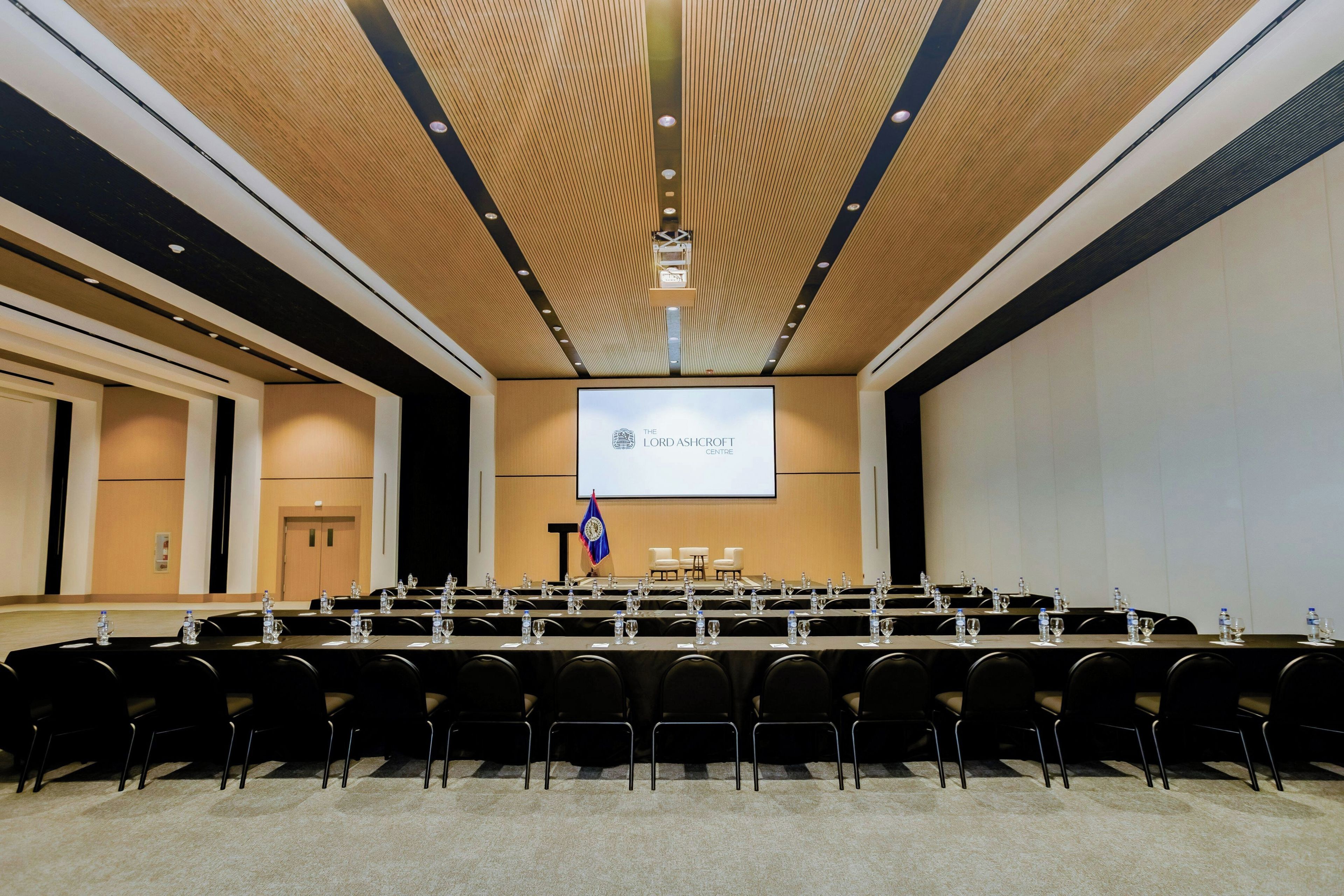 Un salón de conferencias con una pantalla grande mostrando el logotipo del Centro Lord Ashcroft, varias filas de sillas y mesas con botellas de agua.