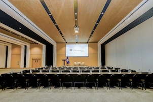 Un salón de conferencias con una pantalla grande mostrando el logotipo del Centro Lord Ashcroft, varias filas de sillas y mesas con botellas de agua.