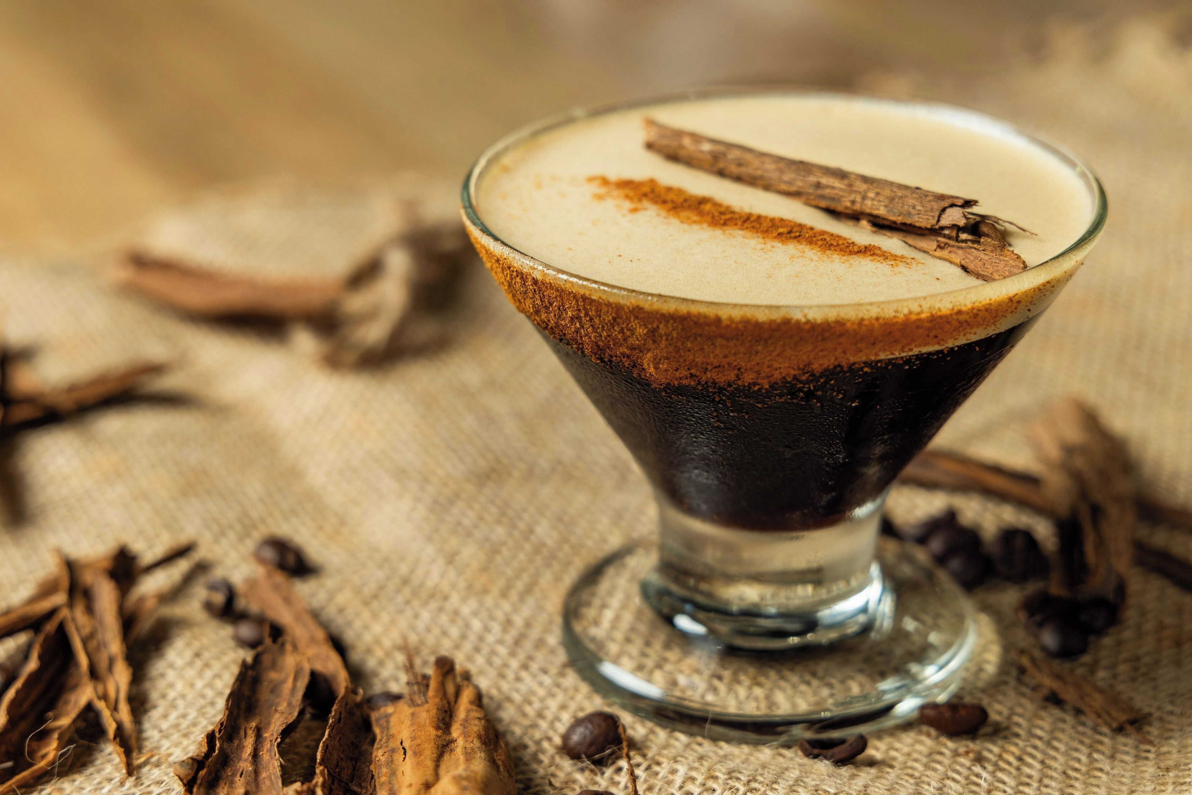 A clear glass cocktail with a layered drink, a cinnamon stick garnish, and coffee beans scattered around.