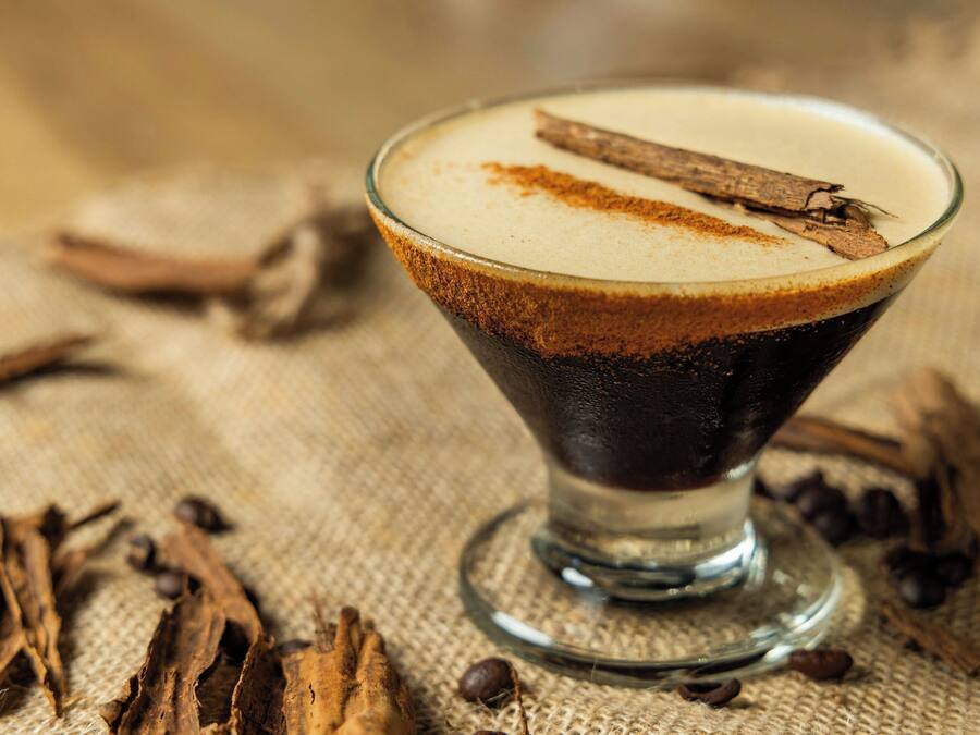 A clear glass cocktail with a layered drink, a cinnamon stick garnish, and coffee beans scattered around.