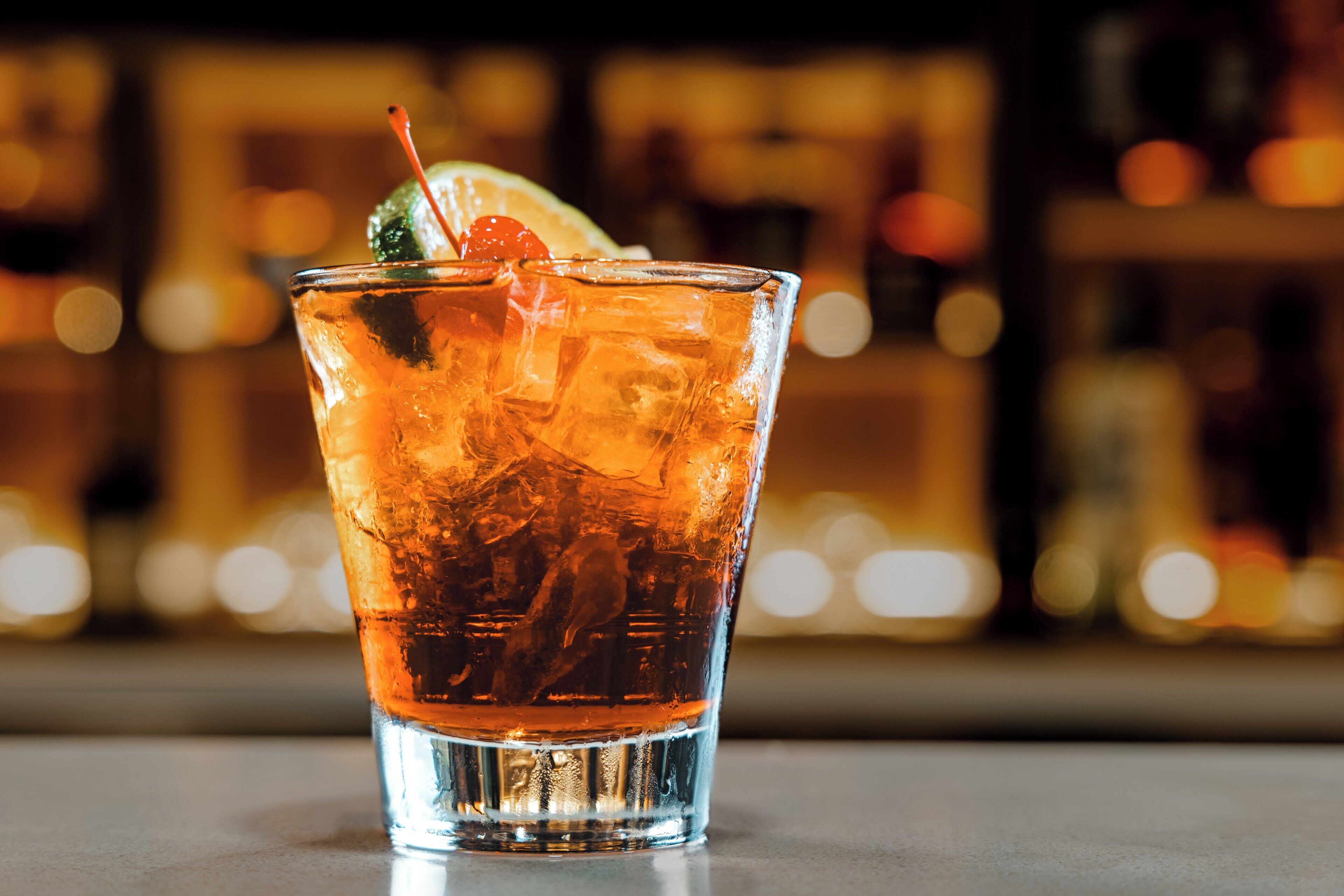 A glass of amber-colored beverage with ice cubes and a garnish, placed on a reflective surface.