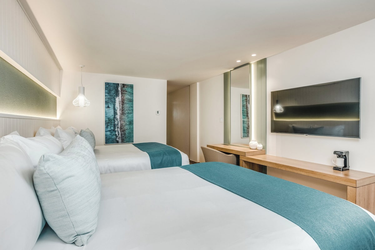 A modern hotel room with two beds, a desk, a flat-screen TV, and a large mirror.