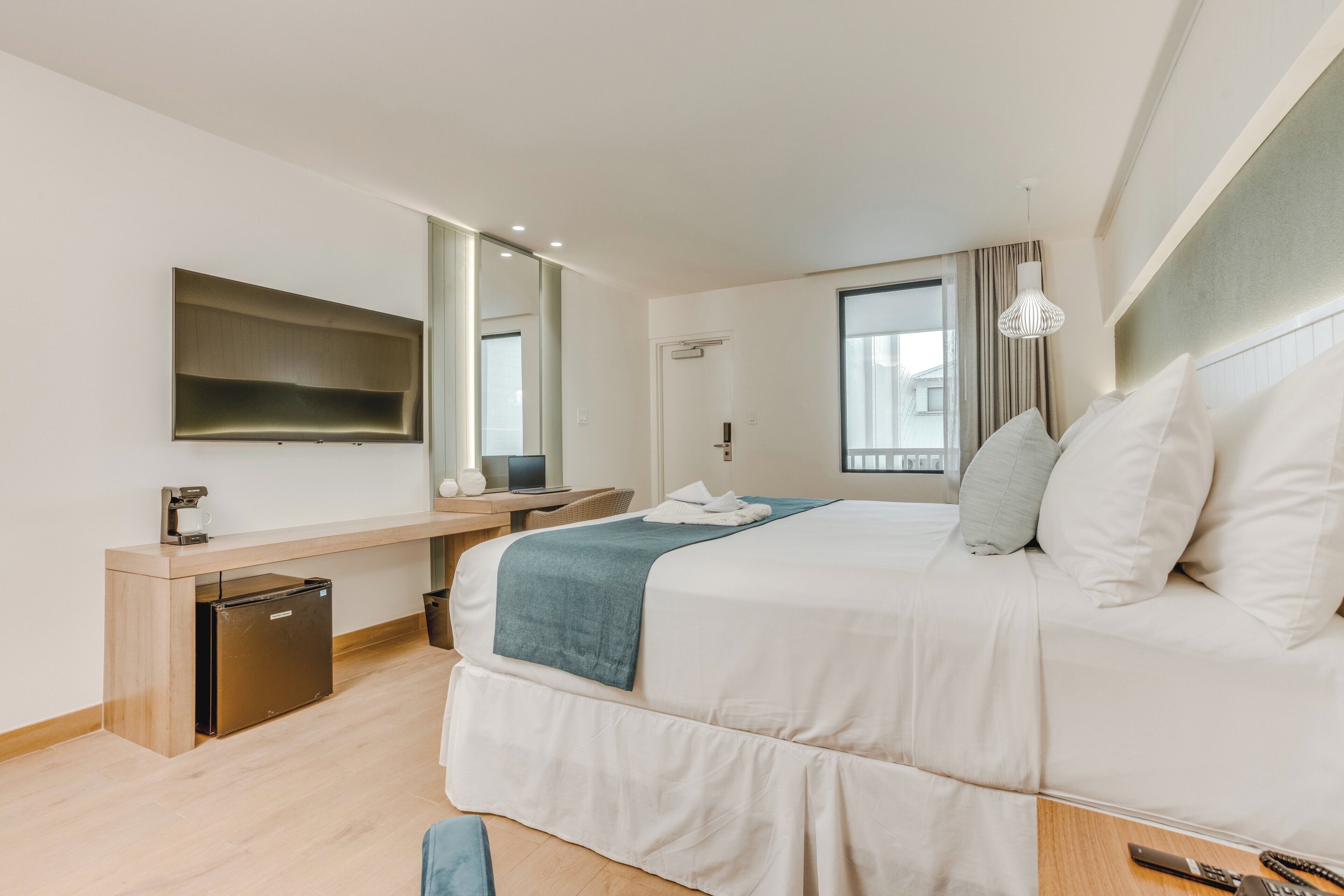 A modern hotel room with a bed, a desk, a mirror, a window, and a small kitchenette.