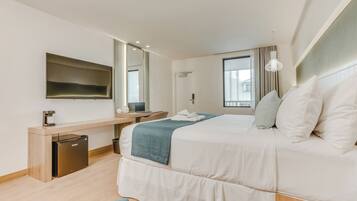 A modern hotel room with a bed, a desk, a mirror, a window, and a small kitchenette.