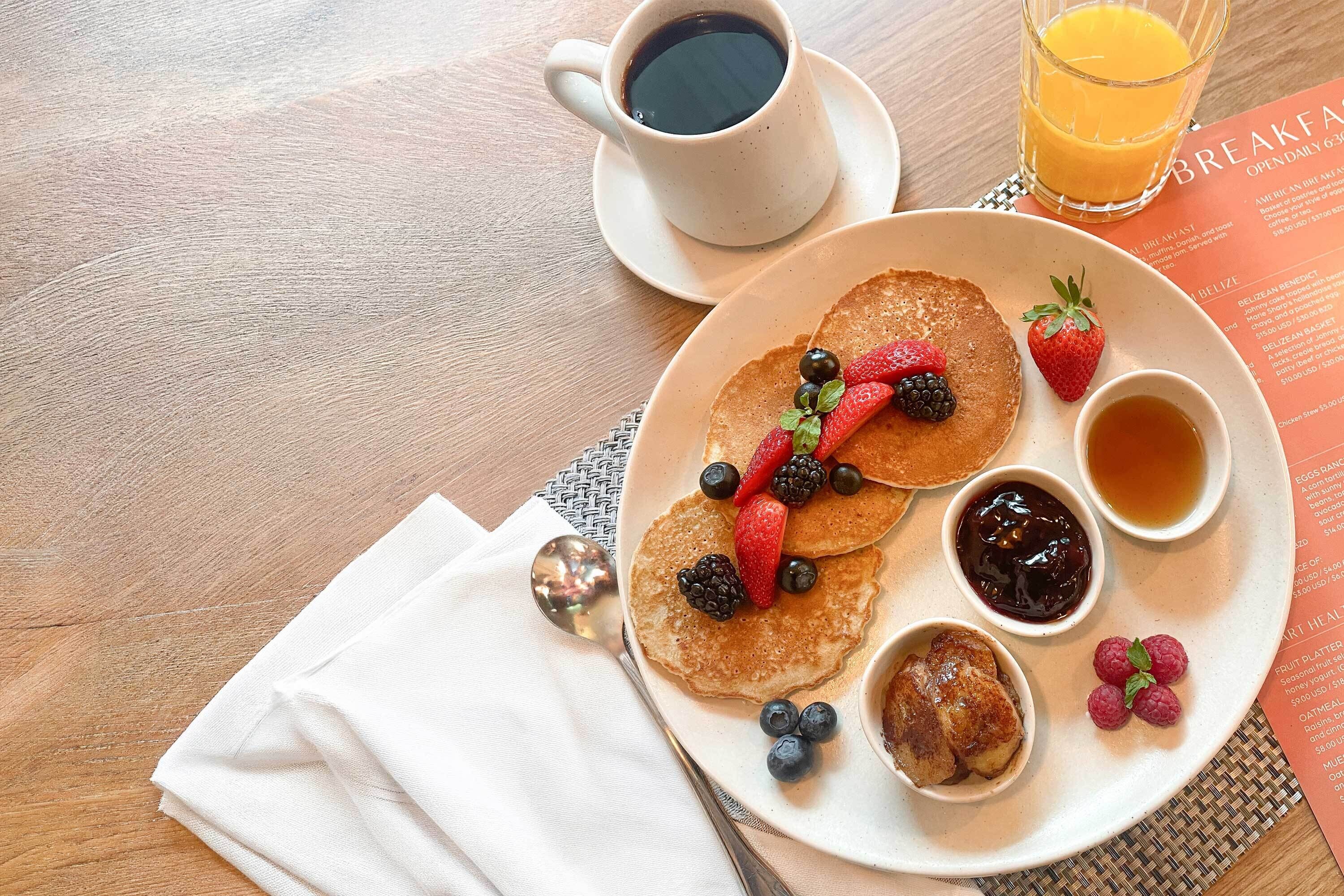 A breakfast plate with pancakes, berries, and syrups, a cup of coffee, and a glass of orange juice on a wooden table.