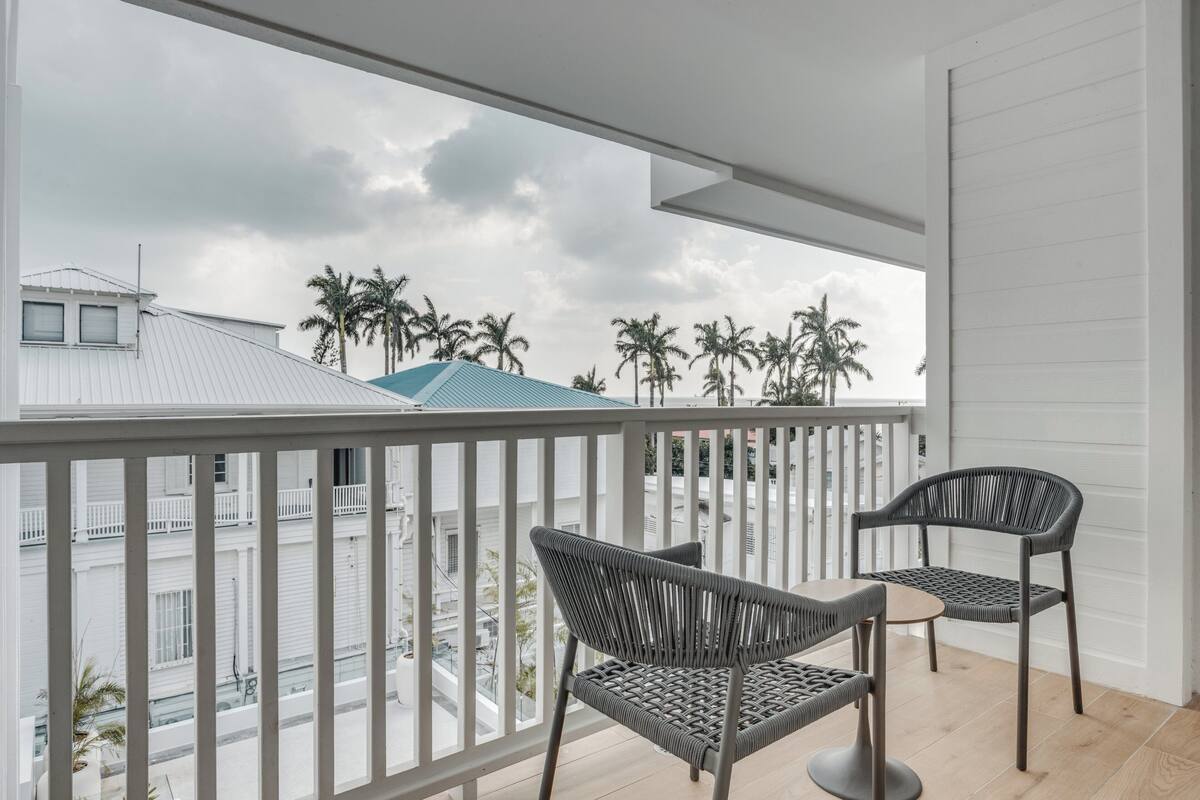 A balcony with two wicker chairs and a small table, overlooking a street with palm trees and buildings.