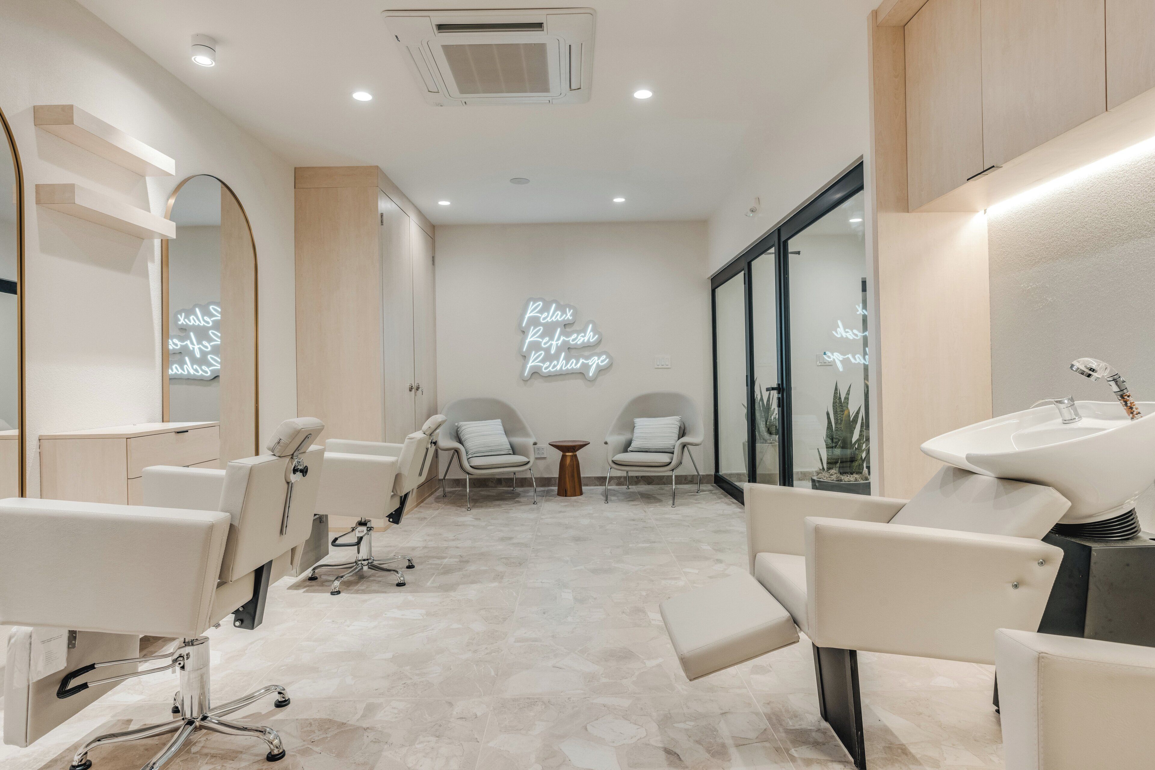 A modern spa room with a large mirror, two chairs, a sink, and a neon sign.