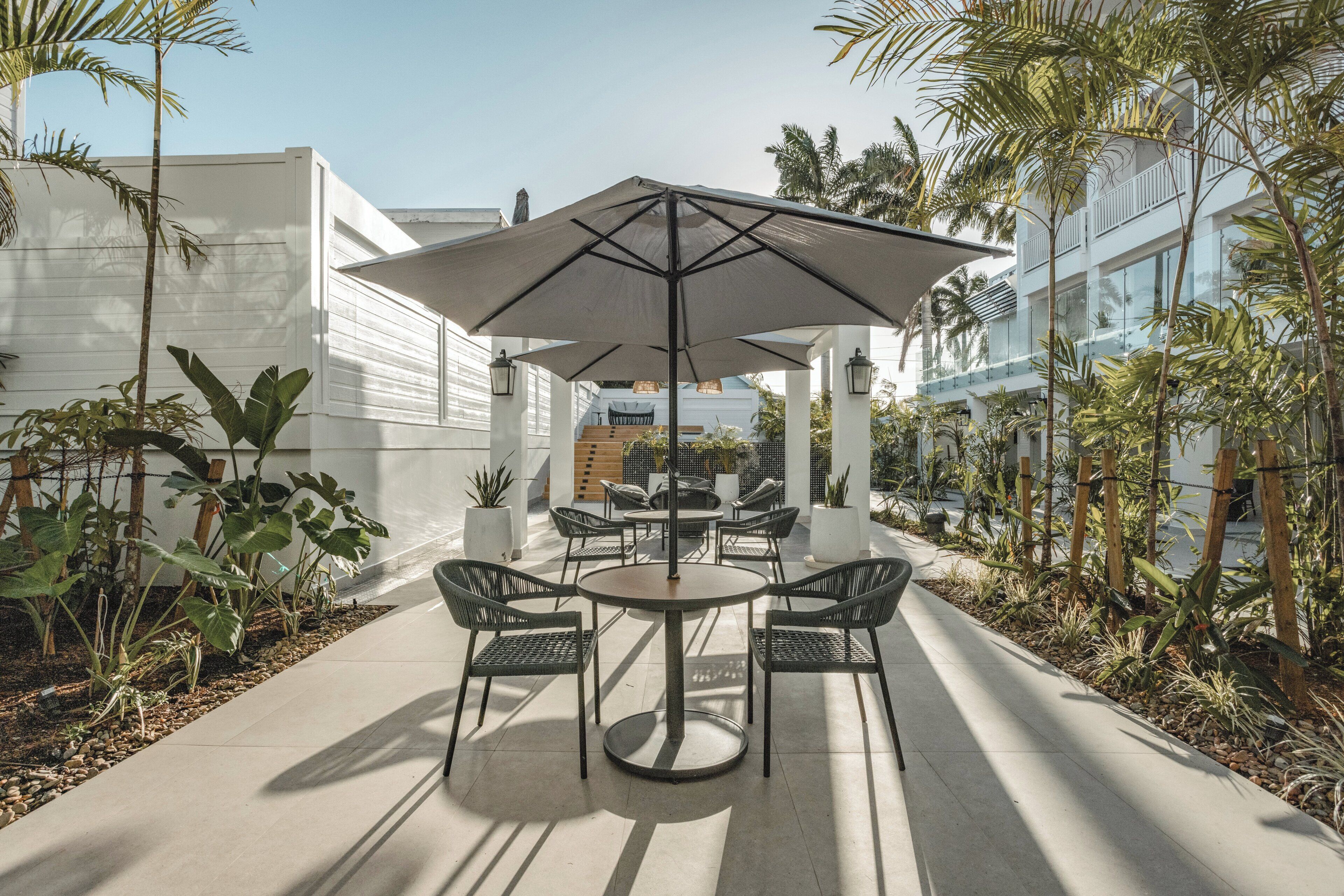 Un patio exterior con una mesa redonda, cuatro sillas, un paraguas grande y plantas en macetas.