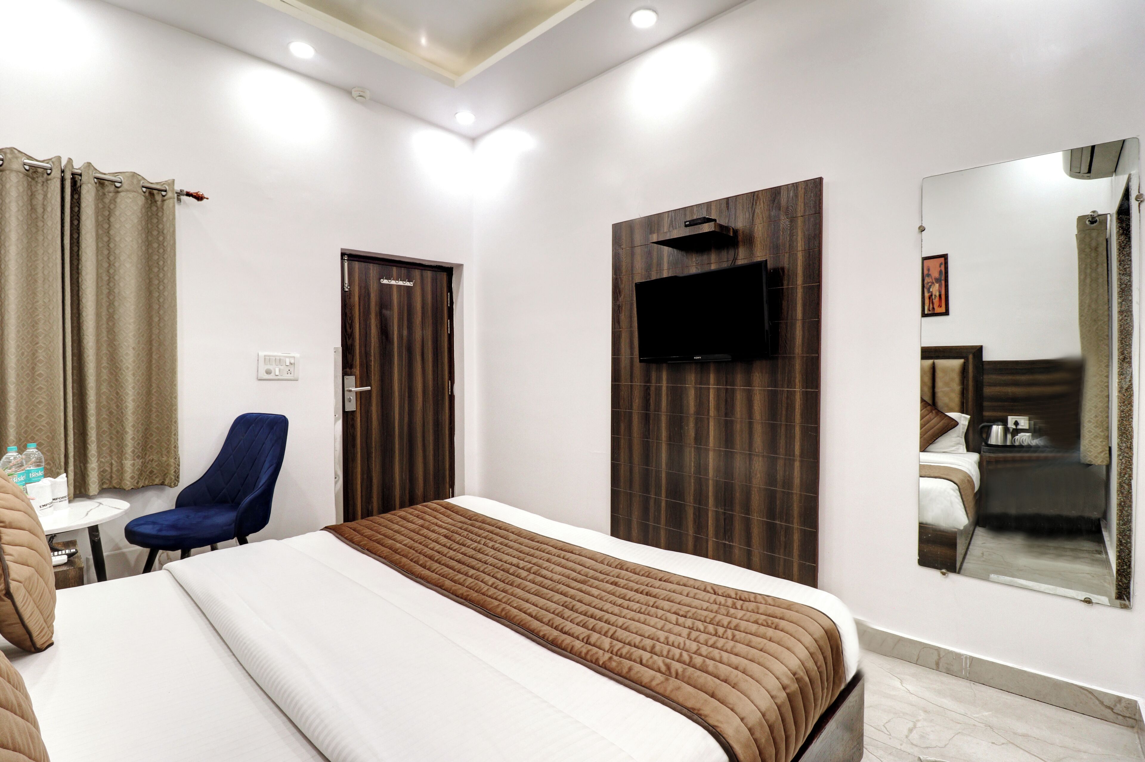 A hotel room with a large bed, a TV mounted on the wall, a chair, a small table with a water bottle, and a door.