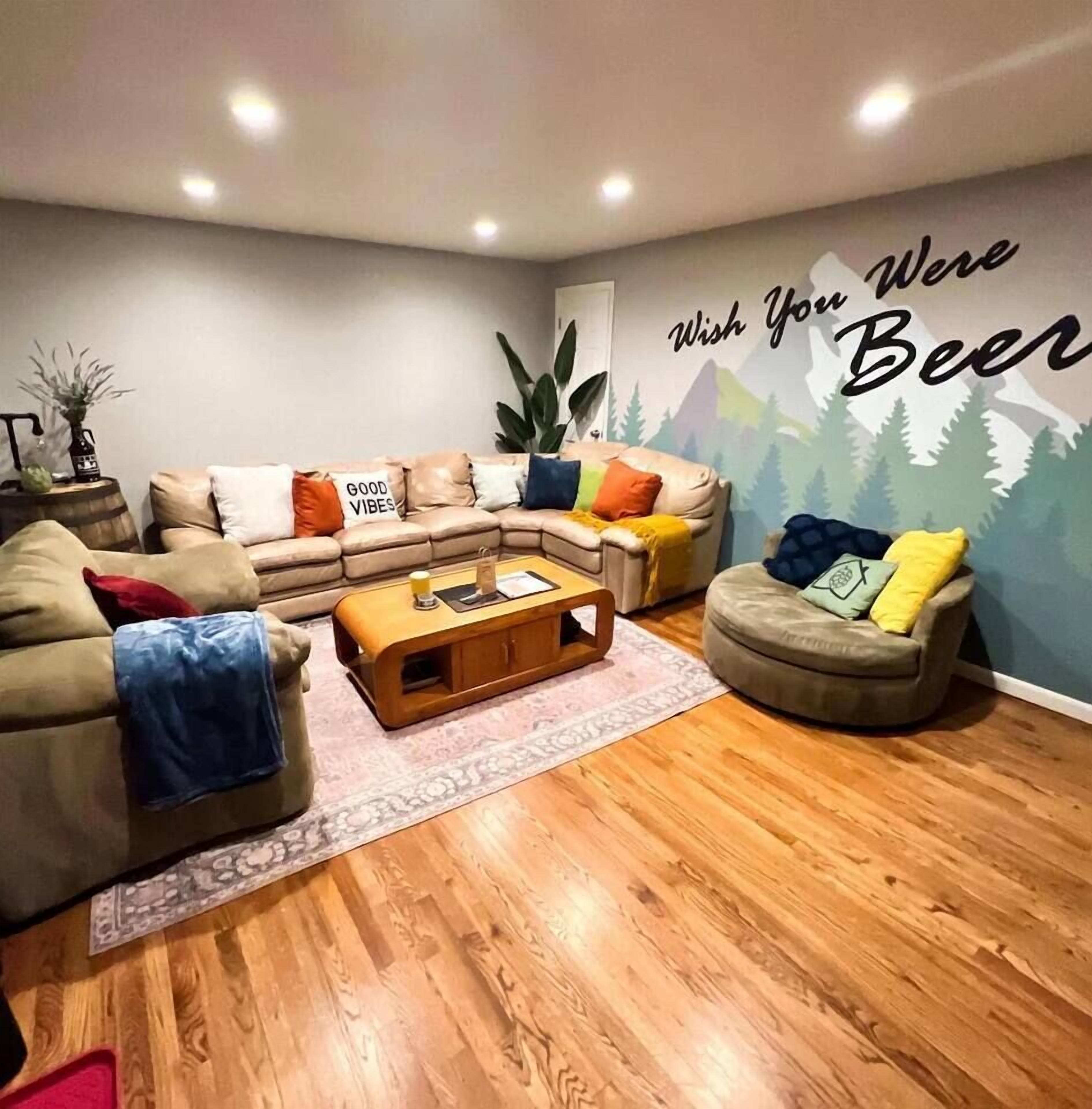 Un salón acogedor con un sofá modular, un reposapiés redondo y una mesa de centro. Un mural reza "Wish You Were Beer".
