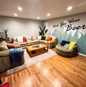 A cozy living room with a sectional sofa, a round ottoman, and a coffee table. A wall mural reads "Wish You Were Beer."