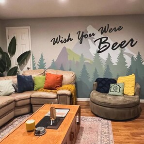 A living room with a sectional sofa, a round ottoman, and a coffee table. There is a wall mural with the text 'Wish You Were Beer.' - Hot Tub, Entertainers Lounge, 6 bedrooms - Denver's Beer Inspired BnB (westminster)