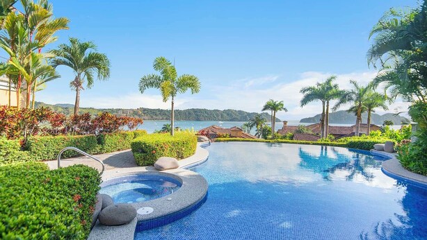 A resort pool with a hot tub, surrounded by tropical plants and palm trees, overlooking a body of water.