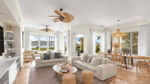 A spacious living room with a large sofa, a coffee table, and a dining area with a wooden table and chairs. There are ceiling fans, large windows, and a view of the outdoors.