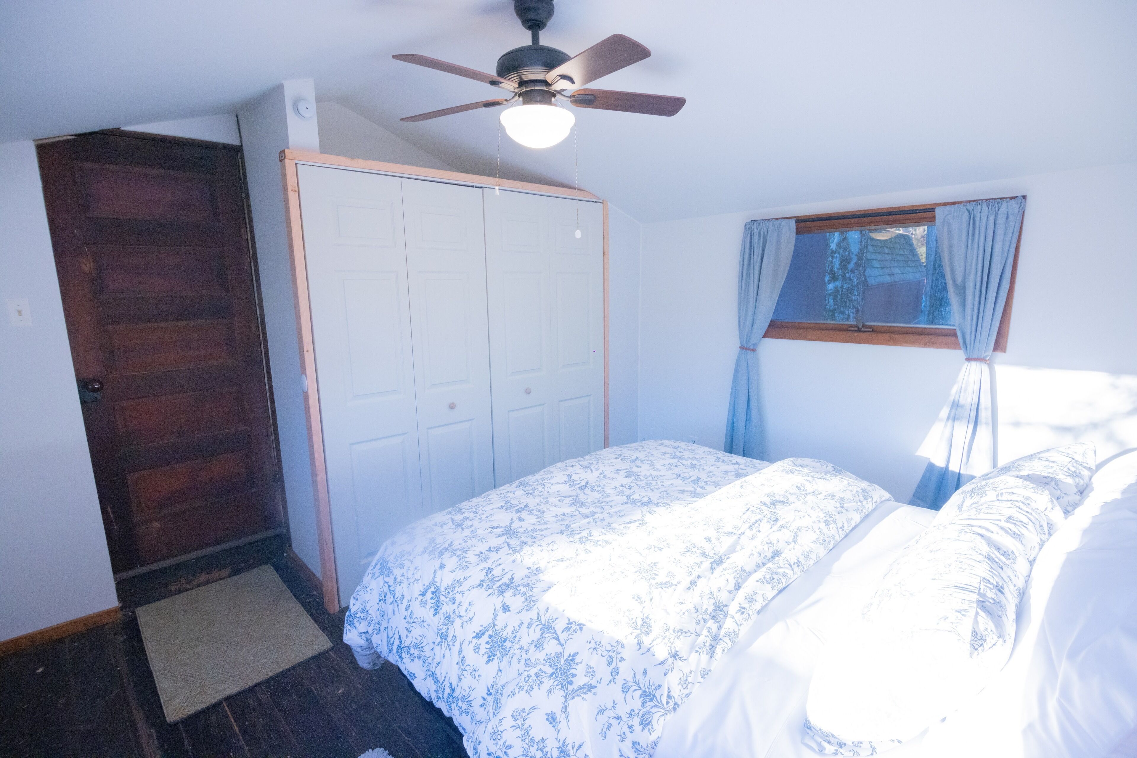 A bedroom with a bed, a ceiling fan, a wardrobe, and a window with curtains.