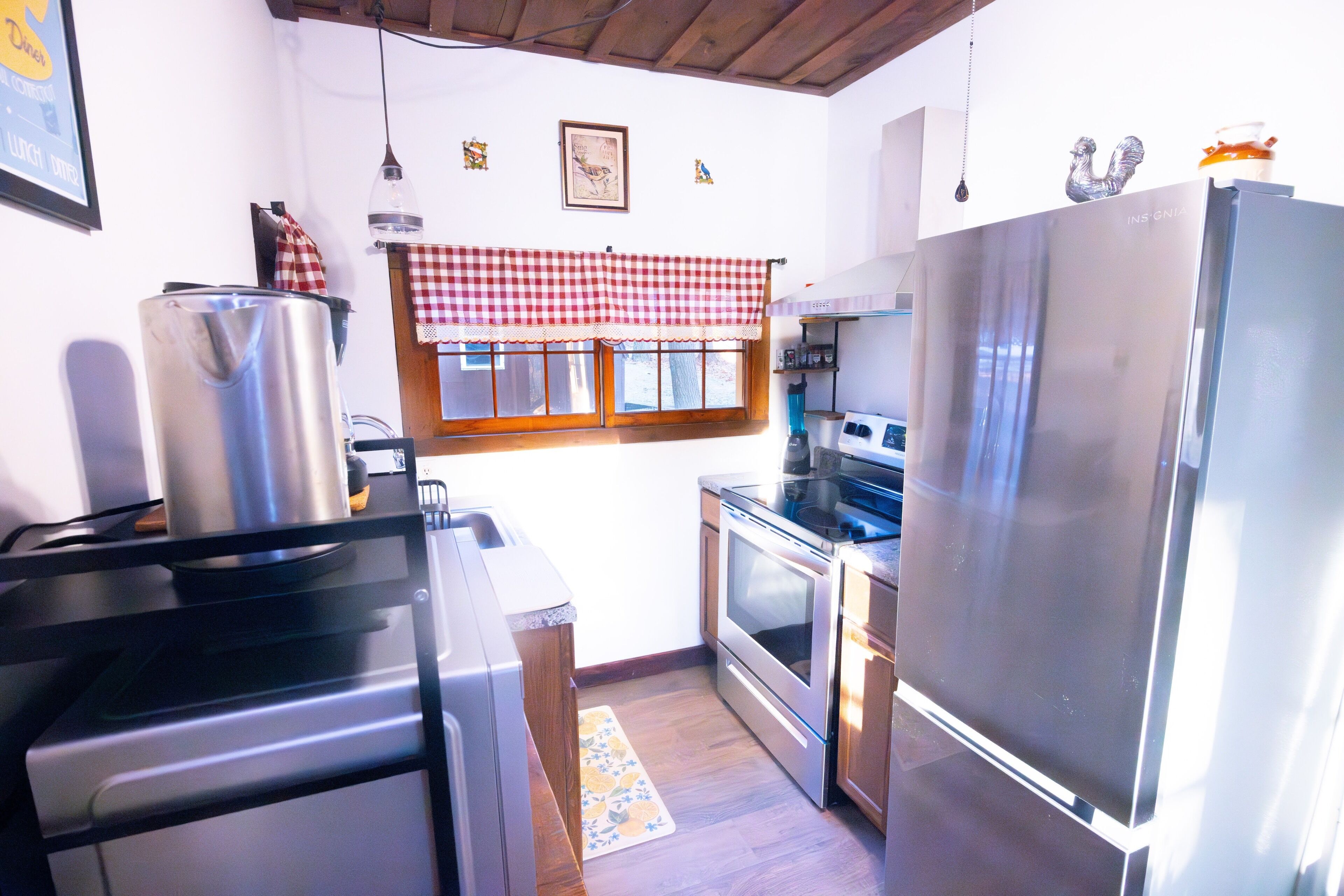 A compact kitchen with stainless steel appliances, a checkered window valance, and a wooden ceiling.