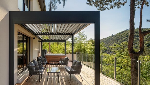Eine Holzterrasse mit Sitzgelegenheiten im Freien, einer Pergola und Blick auf einen bewaldeten Hang.