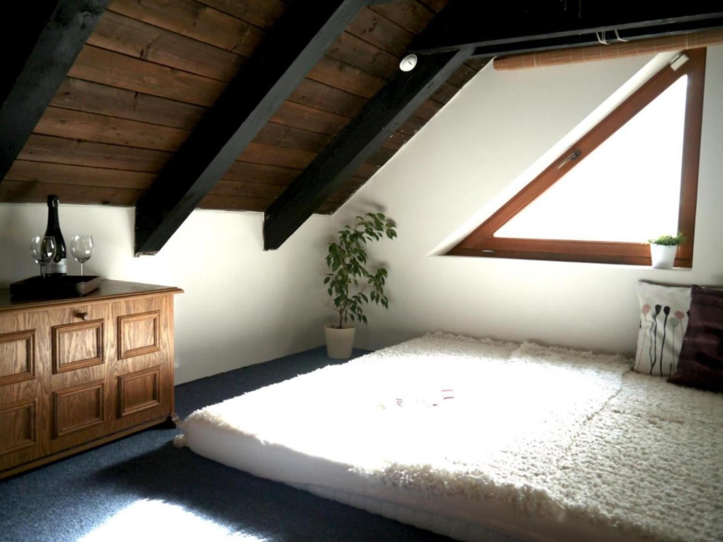 A room with a sloped ceiling, a wooden dresser, a potted plant, a window, and a mattress on the floor.