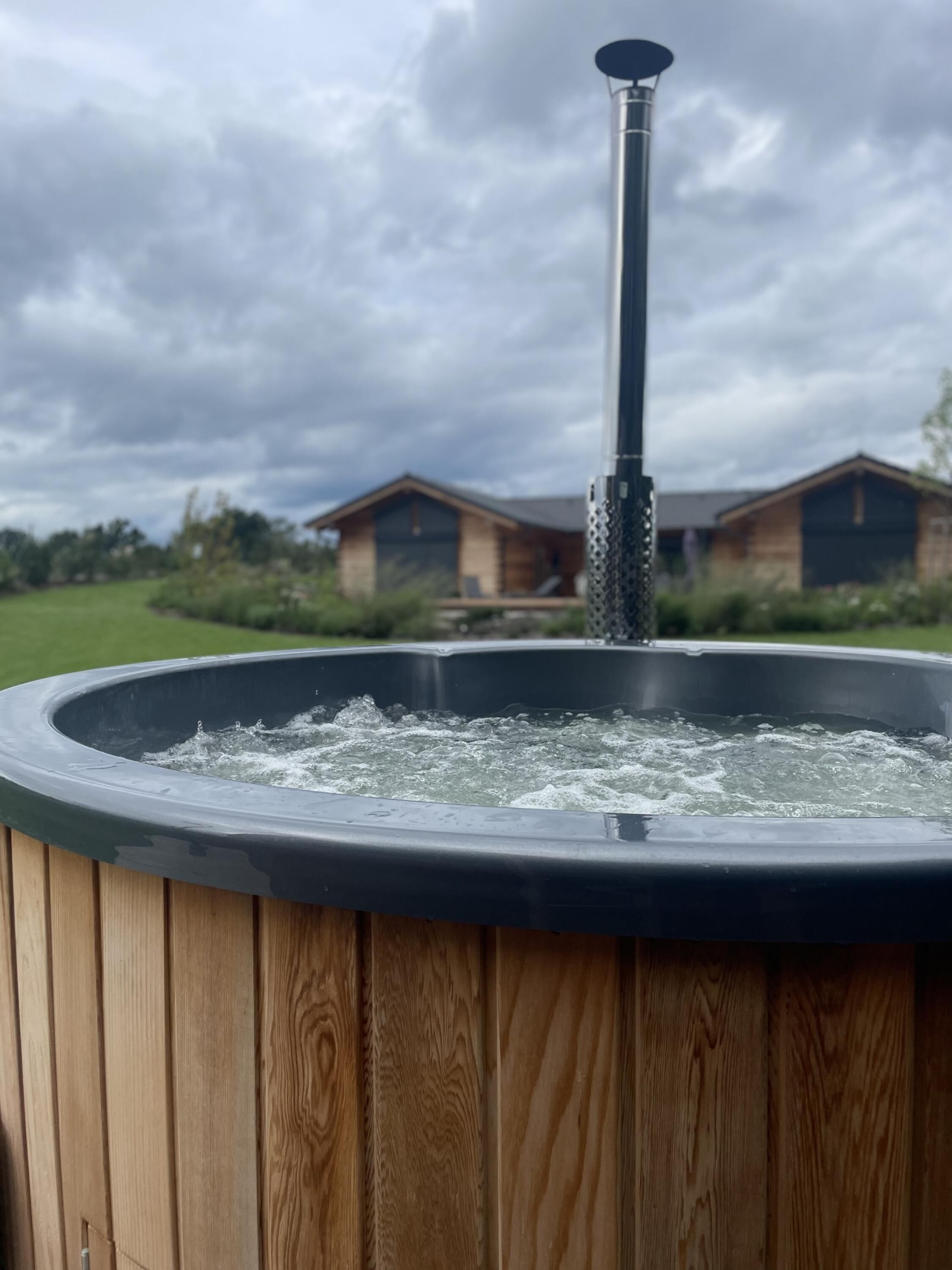 Un jacuzzi con revestimiento de madera, frente a dos cabañas de madera bajo un cielo nublado.