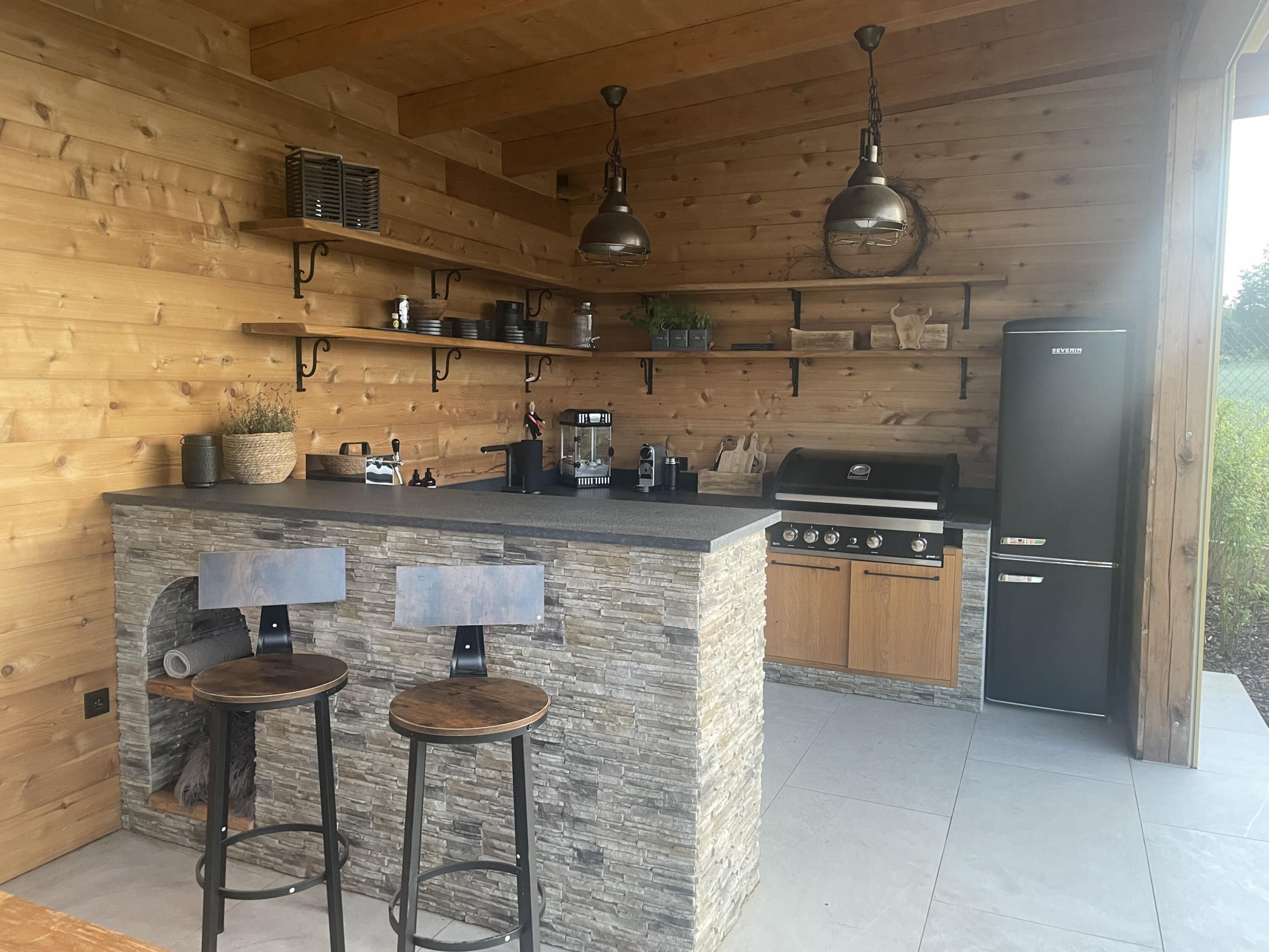 A wooden cabin-style kitchen with a stone island, bar stools, a grill, and a refrigerator.