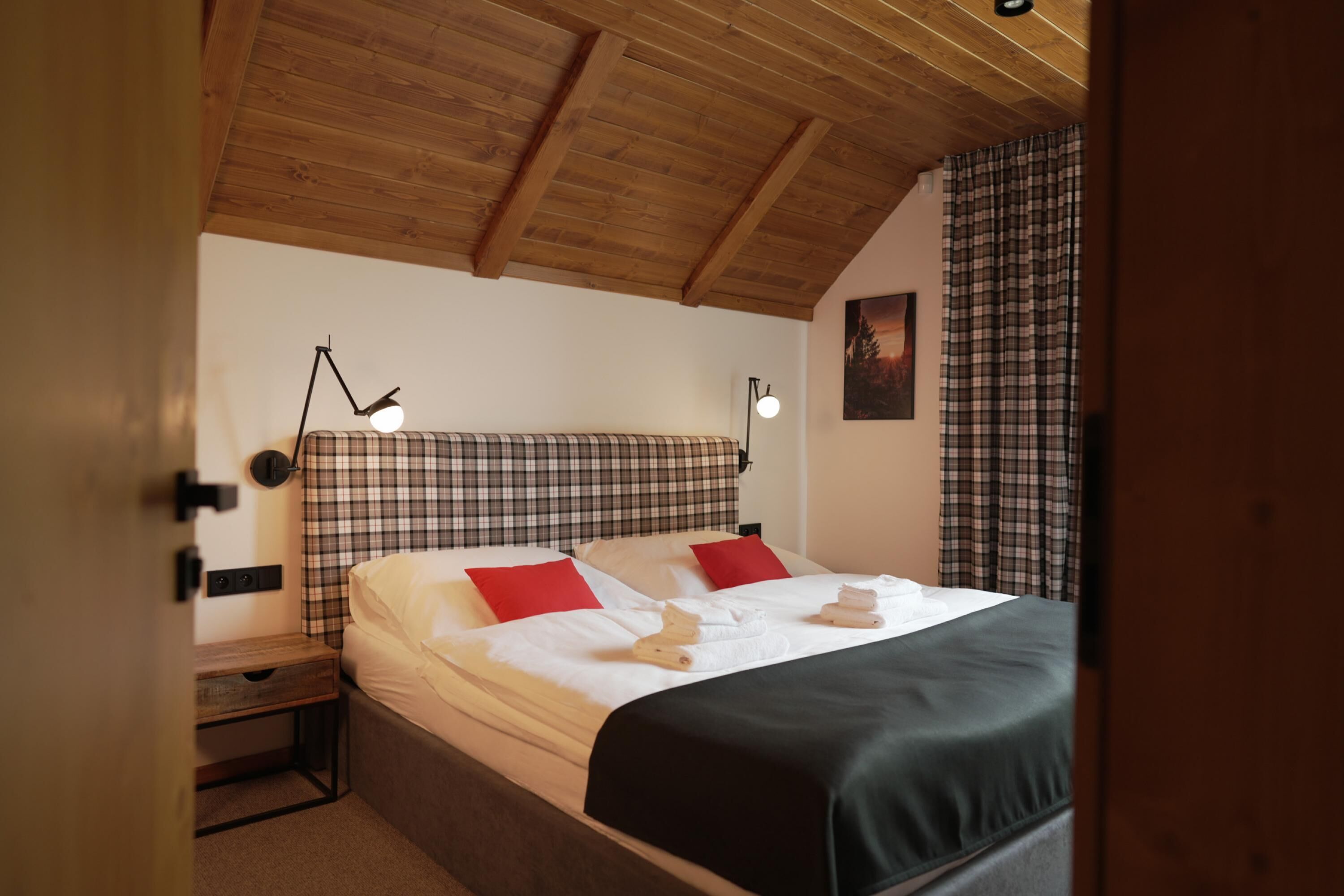 A bedroom with a wooden ceiling, a bed with white linens, red pillows, and a black throw blanket. There is a nightstand with a lamp and a small wooden box.