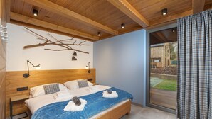 A modern bedroom with a wooden headboard, a large bed with blue bedding, and a view of a garden through a sliding door. - Cerny Dul Kcml115 in Cern Dul (Cerny Dul)