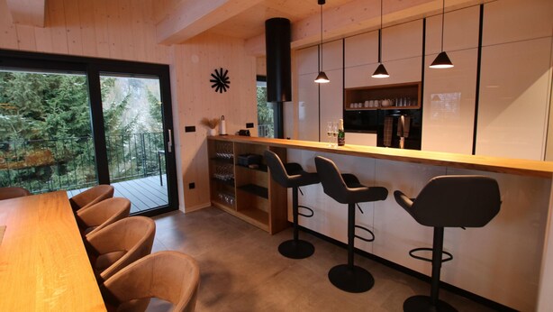 A modern kitchen with wooden cabinetry, a bar area with stools, and a large window overlooking a forested area.