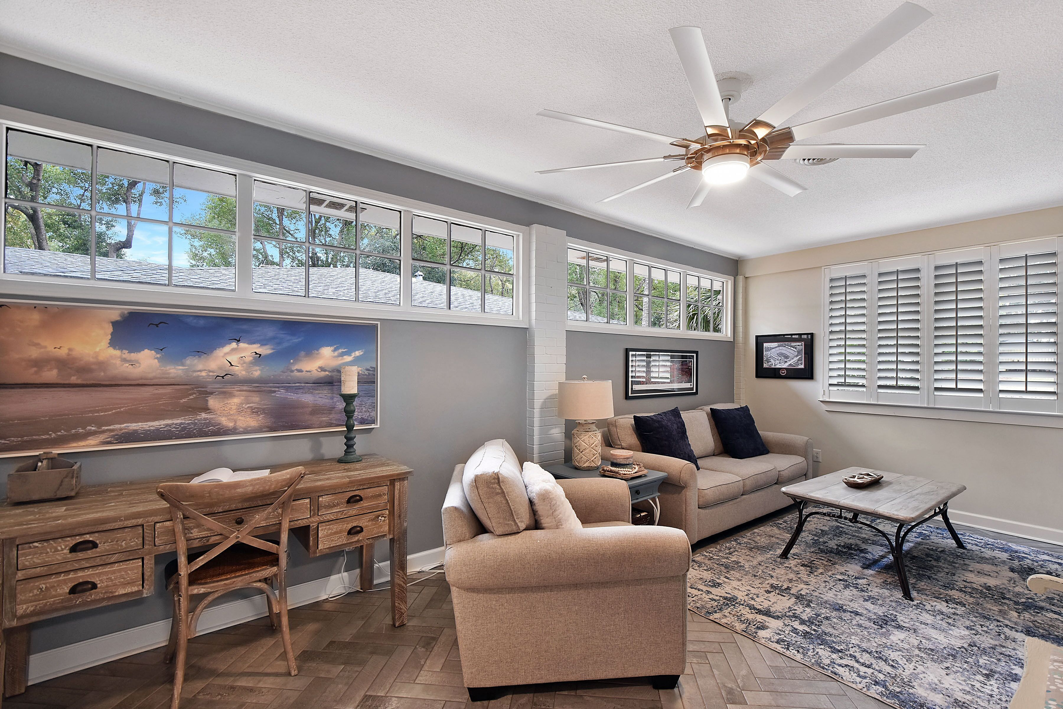 A living room with a large window, a ceiling fan, a sofa, a coffee table, and a desk with a chair.